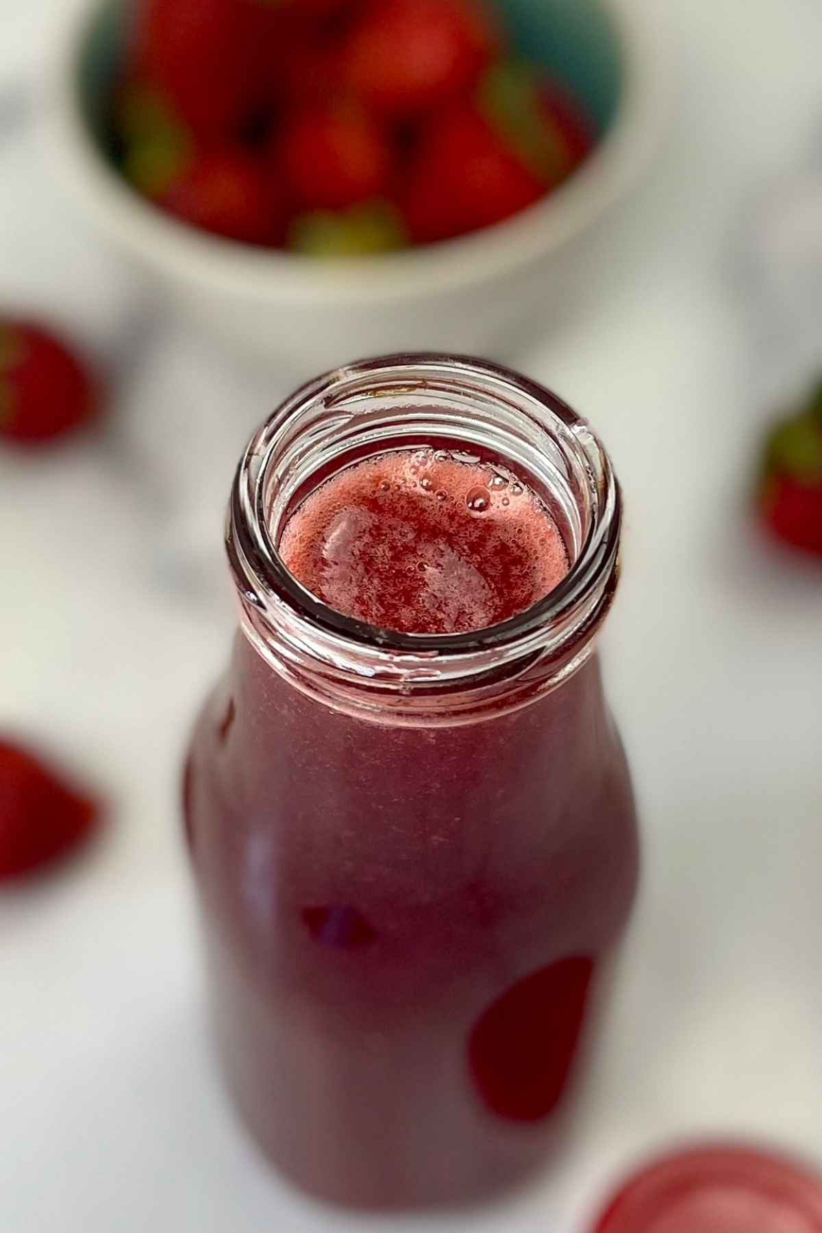 Overhead view of a glass bottle filled with homemade strawberry syrup, with a bowl of strawberries in the background.