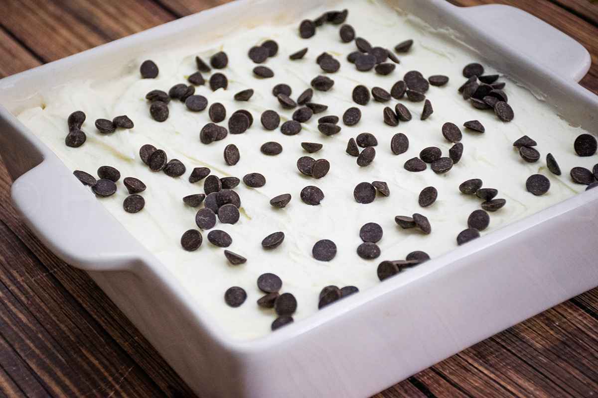 Whole cannoli poke cake in a baking dish topped with ricotta frosting and chocolate chips.