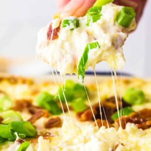 A hand lifting a scoop of gooey baked cheese dip topped with green onions, with melted cheese stretching from the dish.