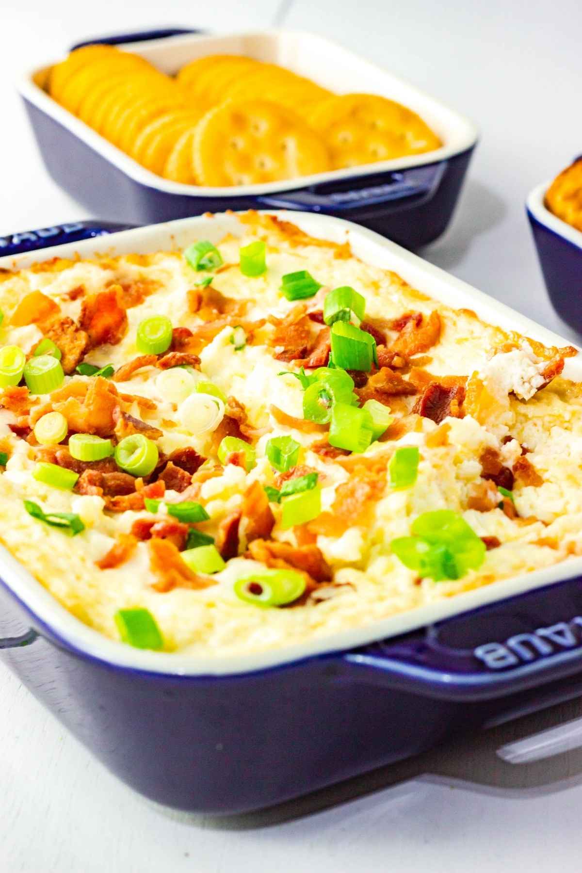 Overhead angled view of a baked cheesy bacon dip topped with fresh green onions and crispy bacon with crackers in the background.