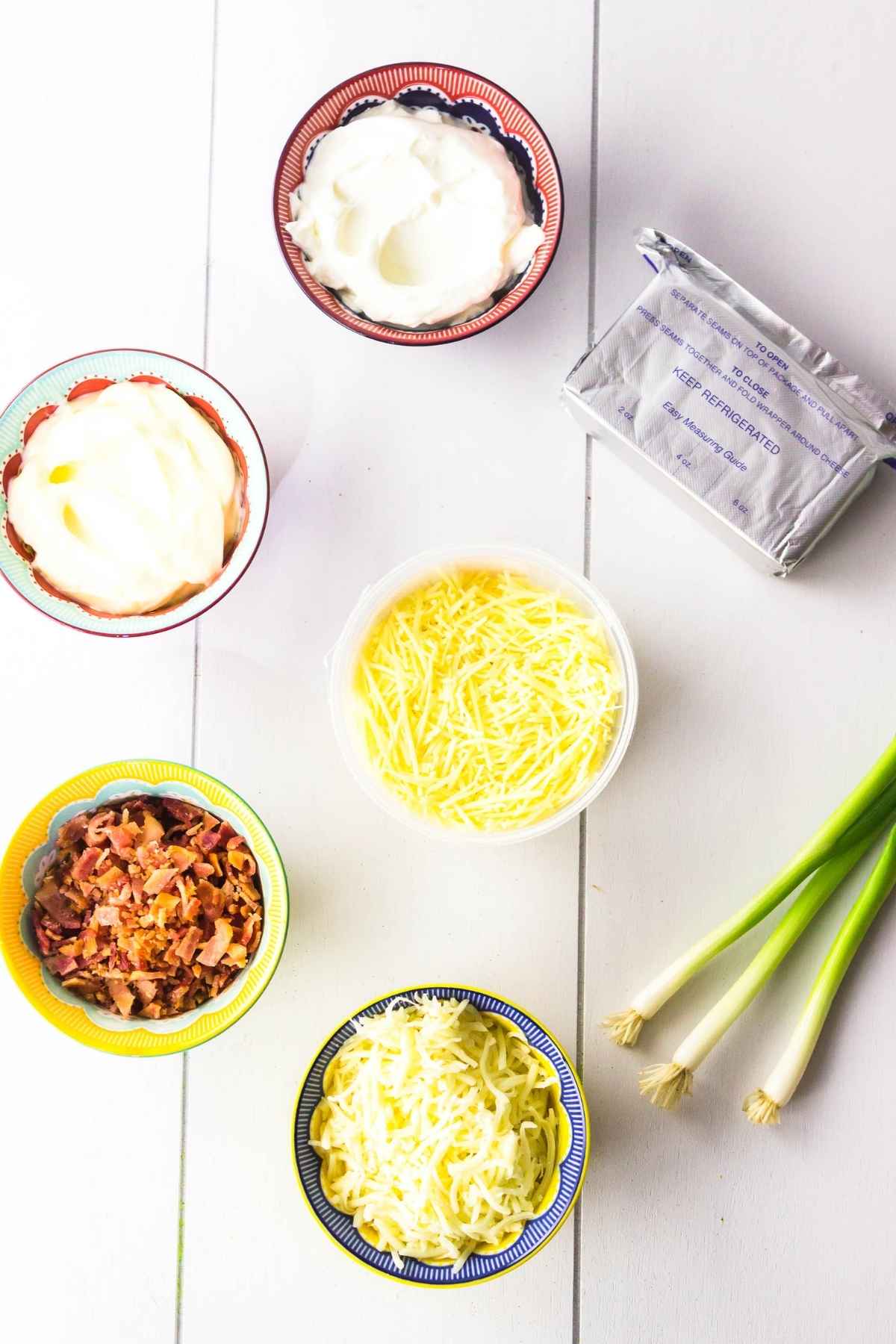 Bowls of shredded cheese, bacon, sour cream, mayonnaise, chopped crispy bacon, cream cheese block, and fresh green onions laid out on a white table.