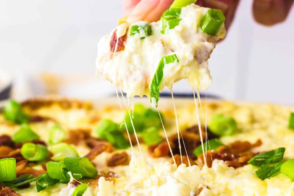 A hand lifting a scoop of gooey baked cheese dip topped with green onions, with melted cheese stretching from the dish.