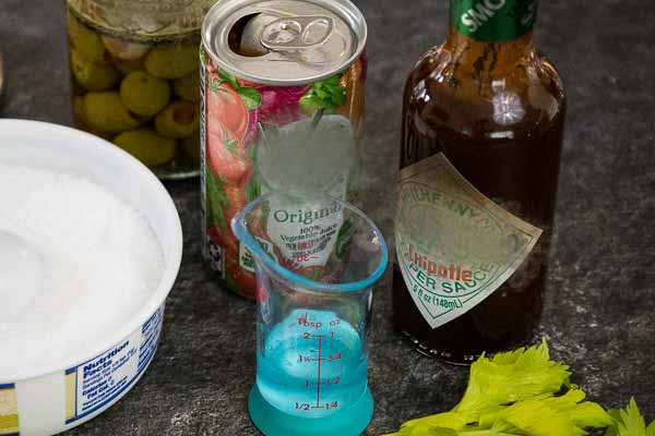 Cocktail ingredients including canned vegetable juice, chipotle hot sauce, a jar of green olives, celery stalks, and a dish of coarse salt.