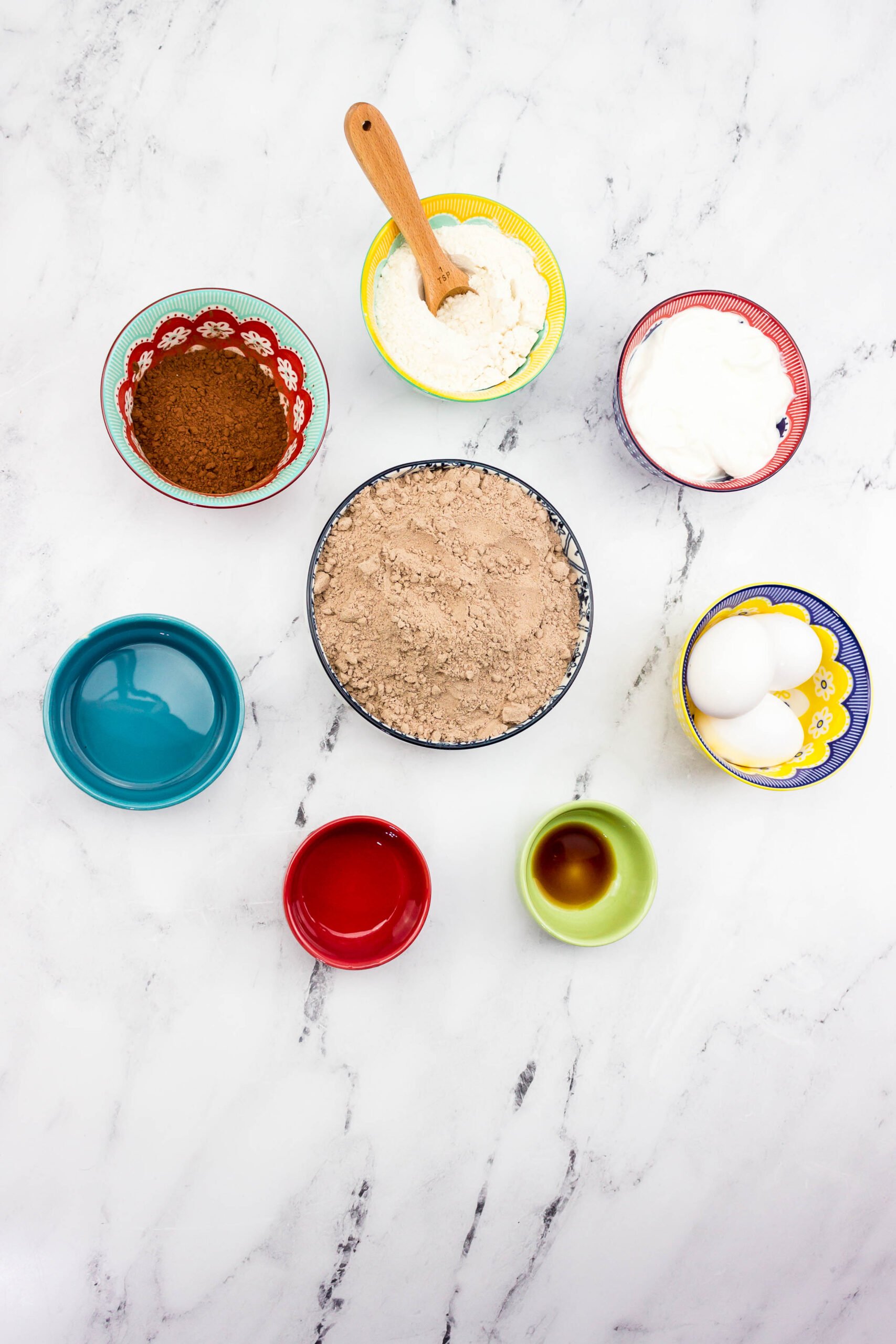Colorful bowls on a marble surface filled with cake mix, cocoa powder, flour, sour cream, eggs, oil, vanilla, and water.