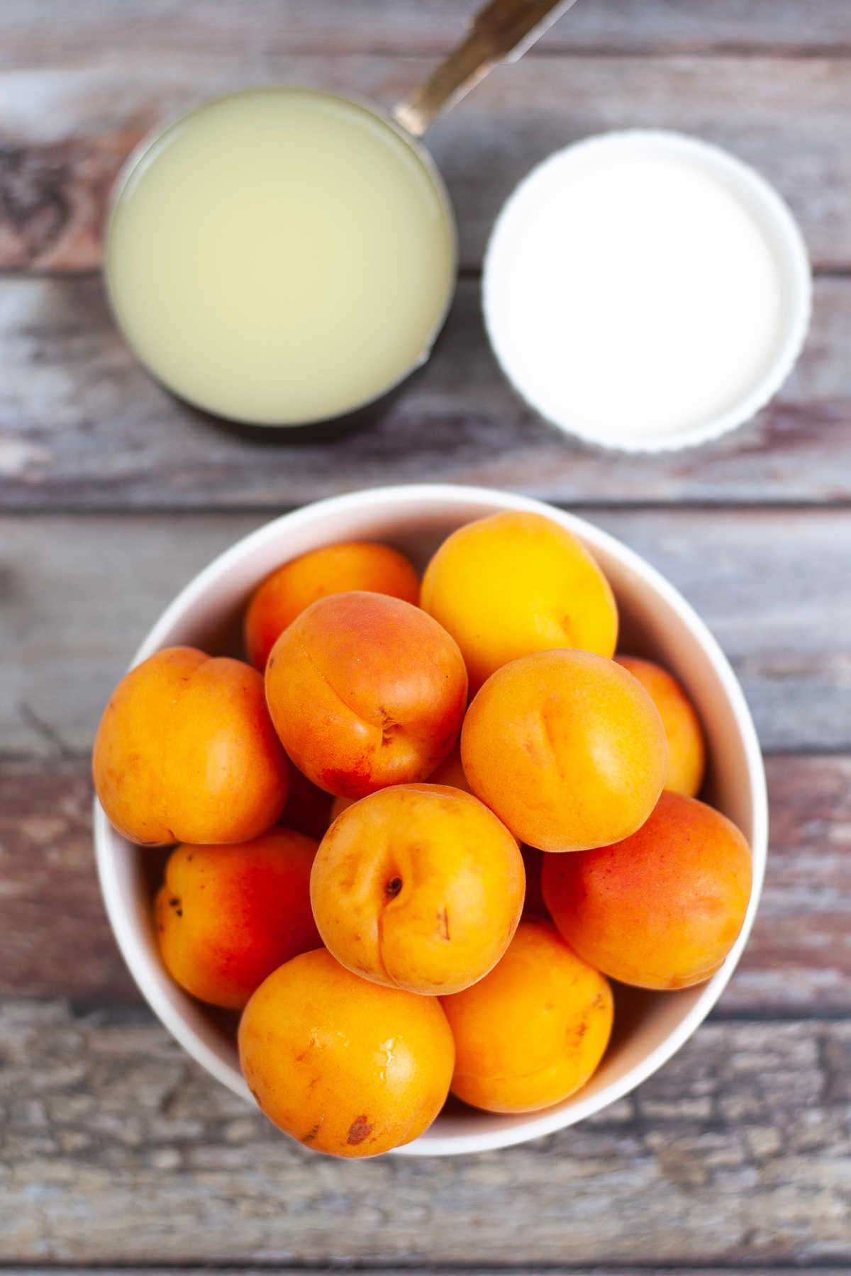 A bowl of fresh apricots with a glass of lemon juice and a small bowl of sugar on a rustic wooden surface.
