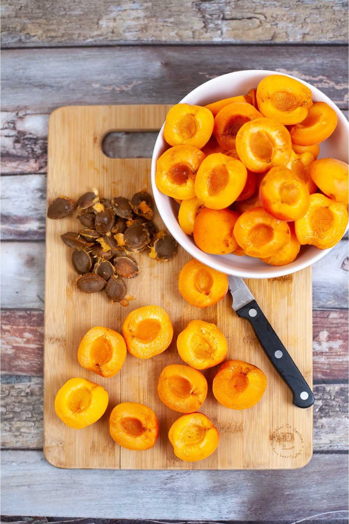 Apricots cut in half with pits removed, sitting on a cutting board next to a paring knife and a pile of seeds.
