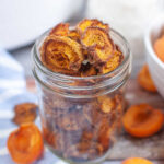 Glass jar filled with dried apricot halves on a rustic table with fresh apricots in the background.