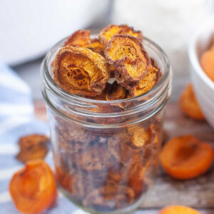 Glass jar filled with dried apricot halves on a rustic table with fresh apricots in the background.