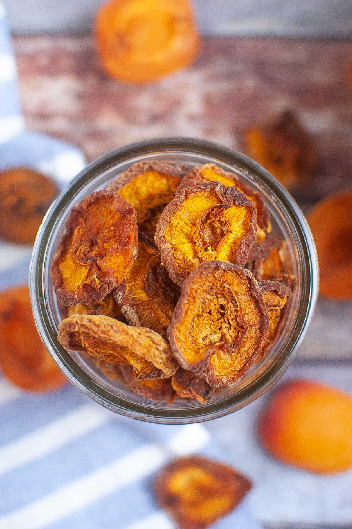 Overhead view of a jar of dried apricots on a striped blue and white cloth with fresh apricots scattered around.