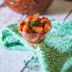 A glass idsh filled with fresh tomato salsa, placed on a rustic white wooden surface with a green patterned cloth.