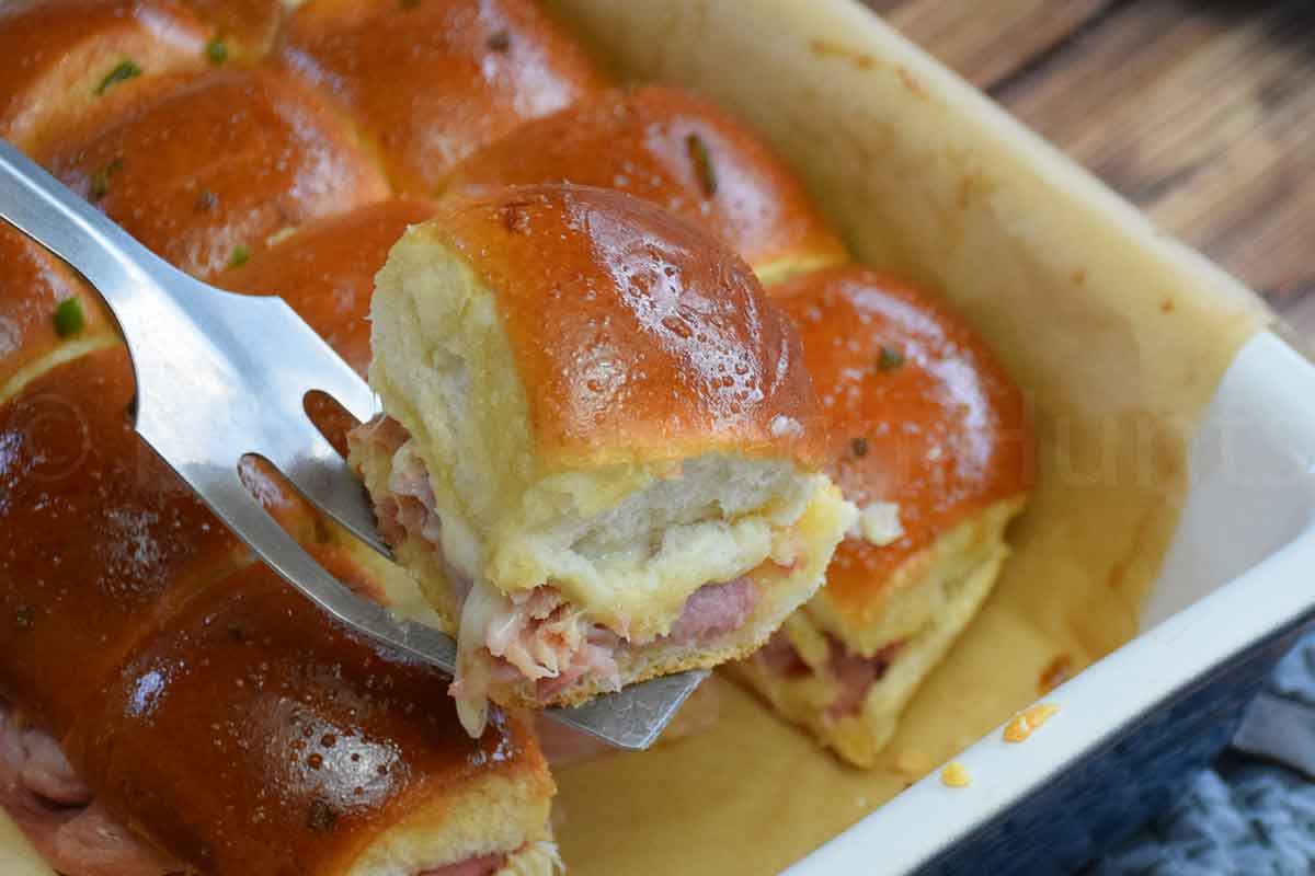 Baked ham and cheese sliders with golden brown tops being lifted from a pn on a metal spatula.