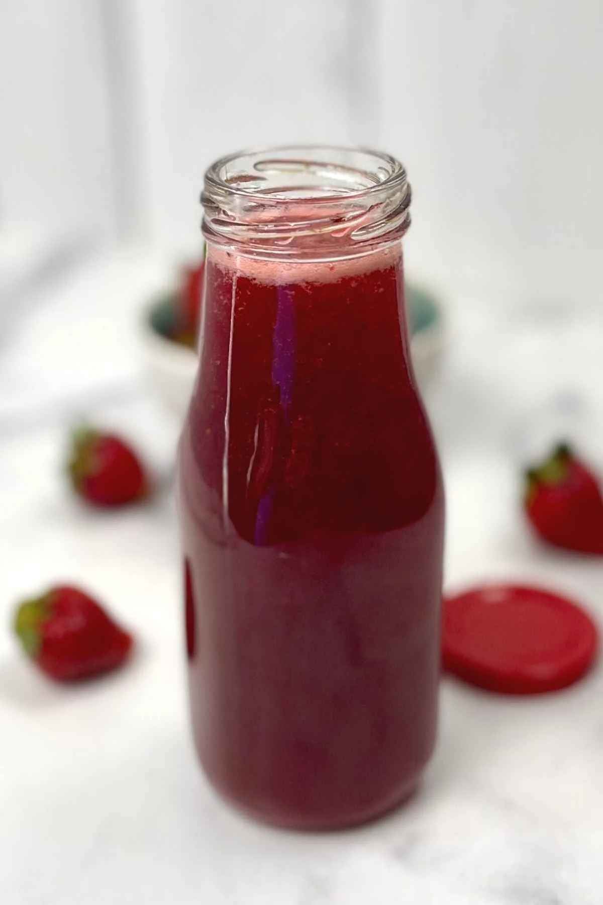 Clear glass bottle filled with homemade strawberry syrup, with fresh strawberries blurred in the background.