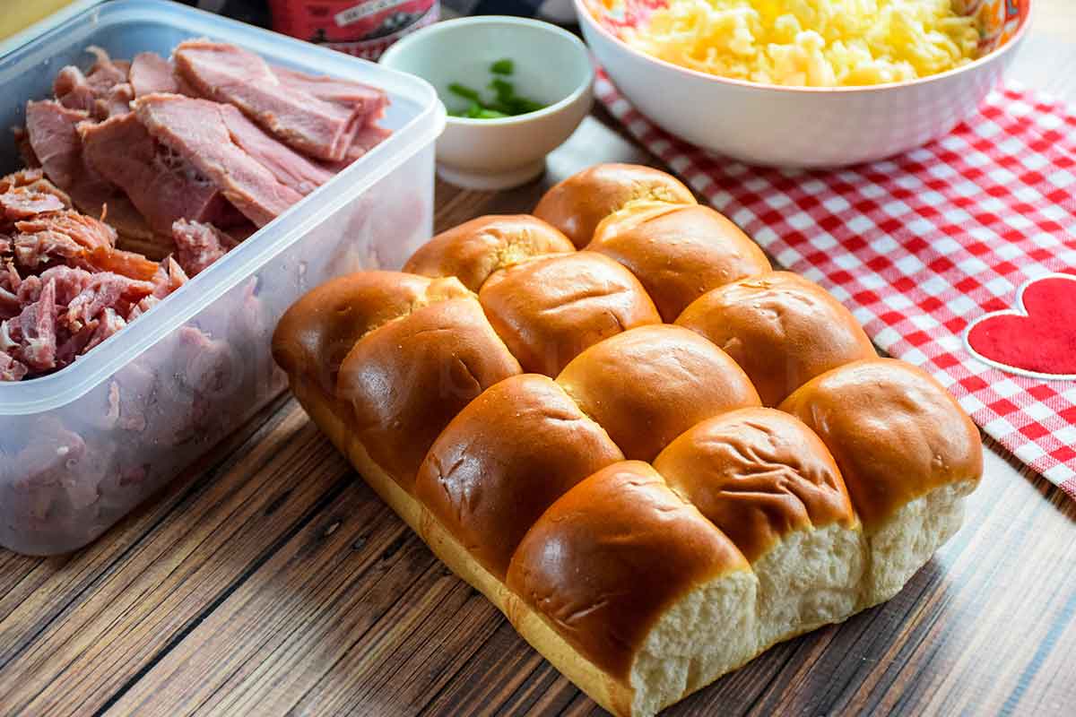 Hawaiian rolls, sliced ham, shredded cheese, and green onions on a wooden table.