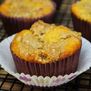 Keto rhubarb muffin with golden brown crumb topping in a purpla paper liner, resting on a cooling rack.