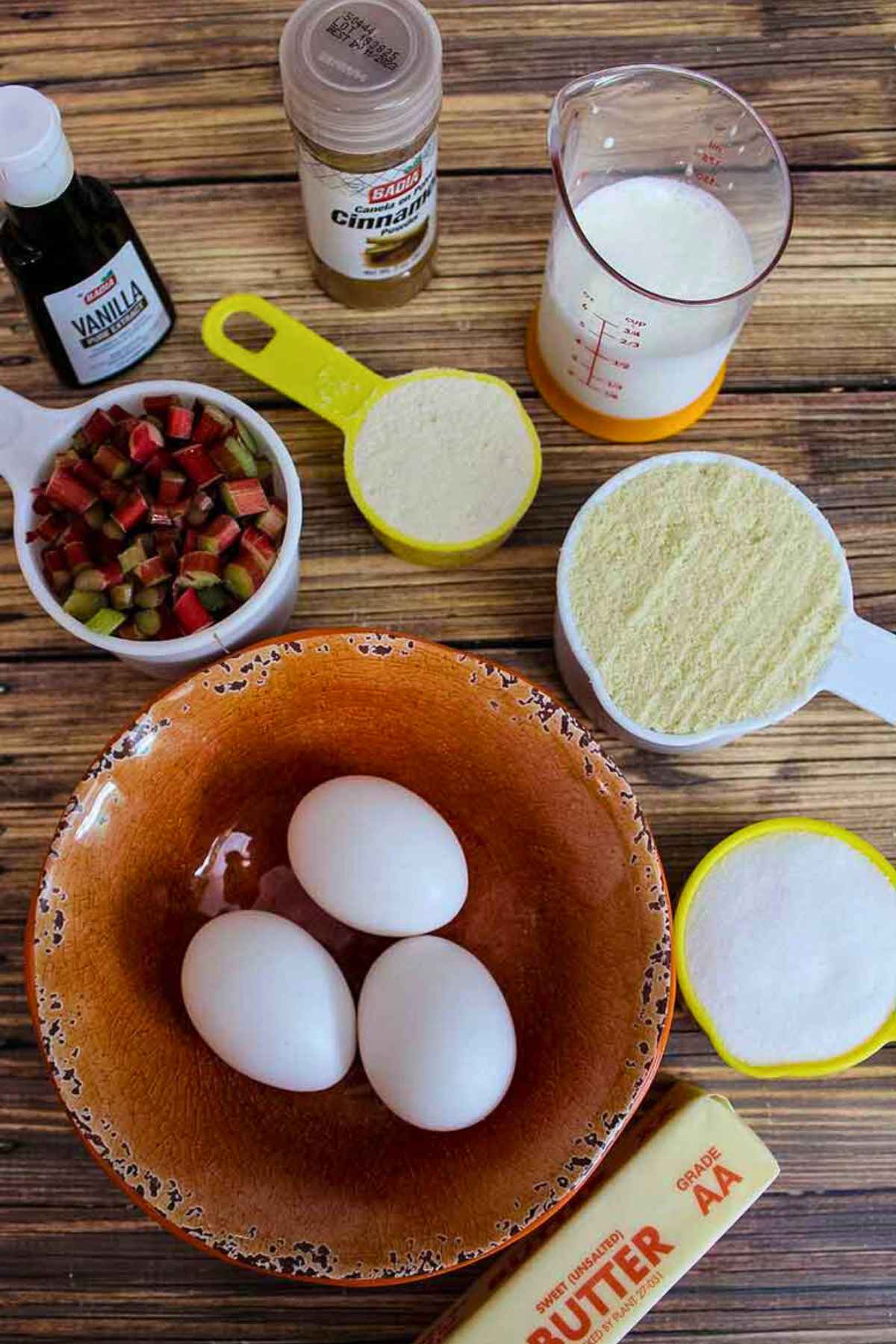 Overhead view of baking ingredients for muffins, including eggs, chopped rhubarb, butter, vanilla, cinnamon, milk, almond flour, and sugar substitute.