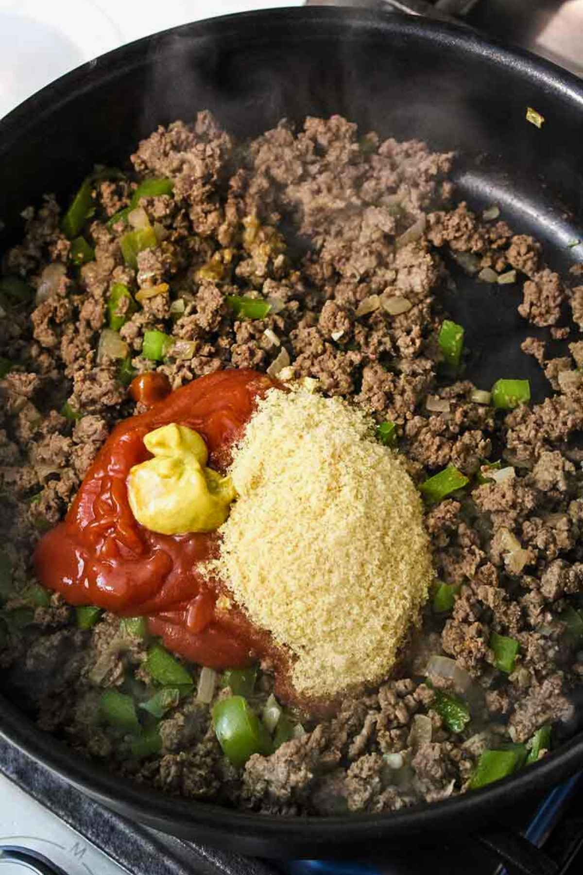 Ground venison and green peppers in a skillet with ketchup, mustard, and seasonings on top before mixing.