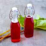 Two glass bottles filled with bright red rhubarb syrup, sealed with swing-top caps, with rhubarb stalks and leaves in the background.