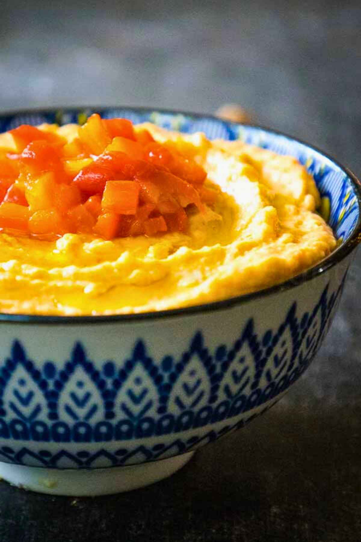 Close up of roasted red pepper hummus in a patterned bowl, garnished with chopped roasted peppers.
