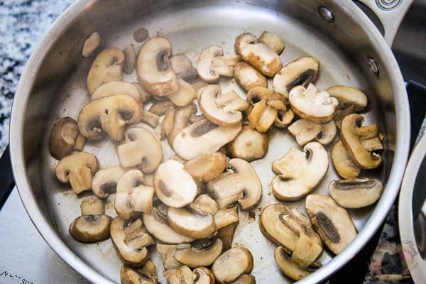 Sliced mushrooms cooking in a stainless steel skillet on a stovetop.