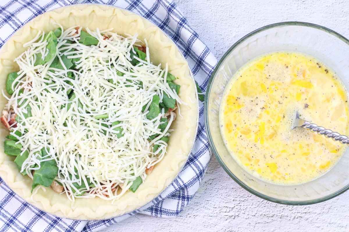 Overhead view of an unbaked quiche filled with cheese and greens in a pie crust, next to a glass bowl of whisked eggs and milk with a spoon.