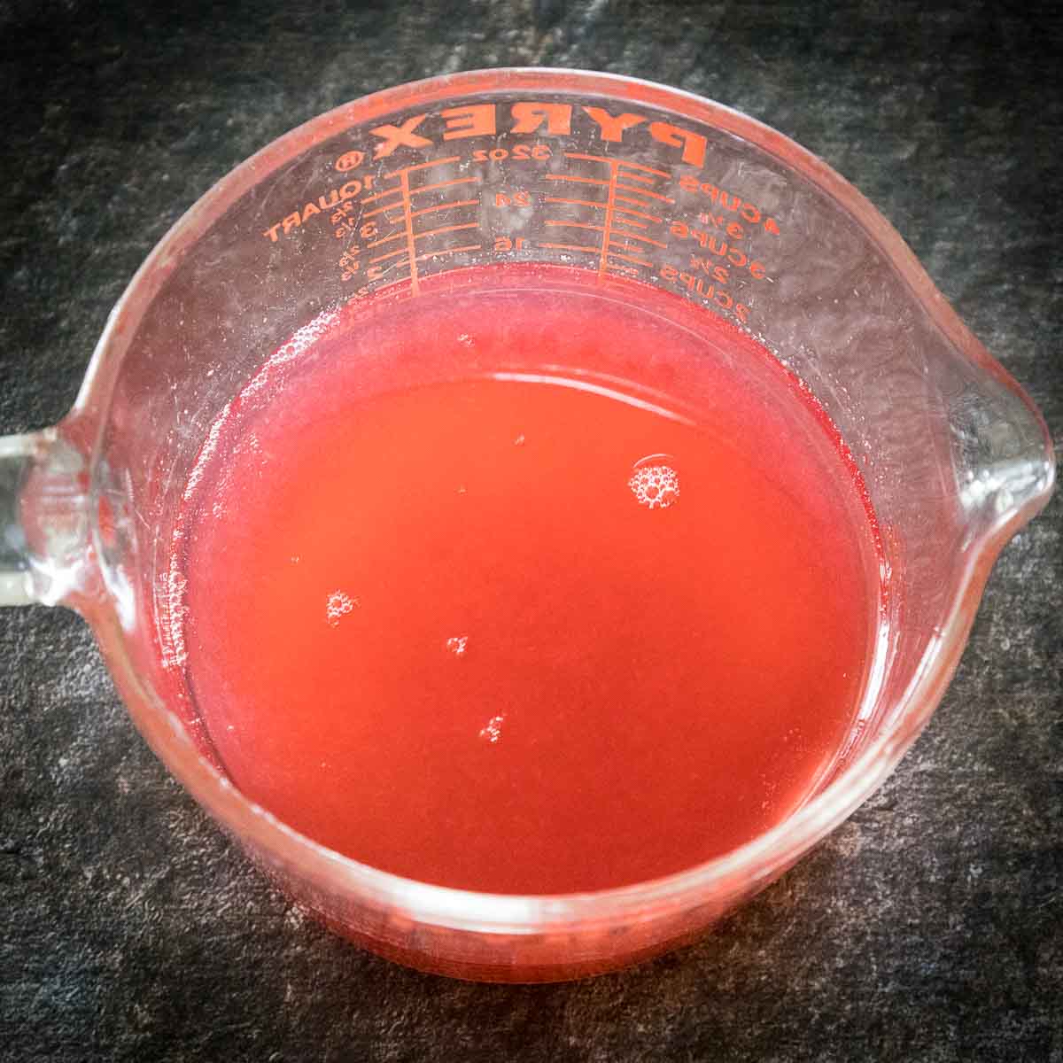 A glass measuring cup filled with strained red rhubarb syrup on a dark surface.