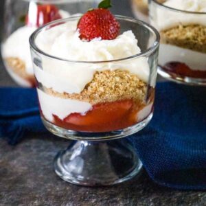 A glass dessert cup fllled with layers of strawberry sauce, creamy cheesecake filling and crushed vanilla wafer cookies topped with whipped cream and a fresh strawberry.