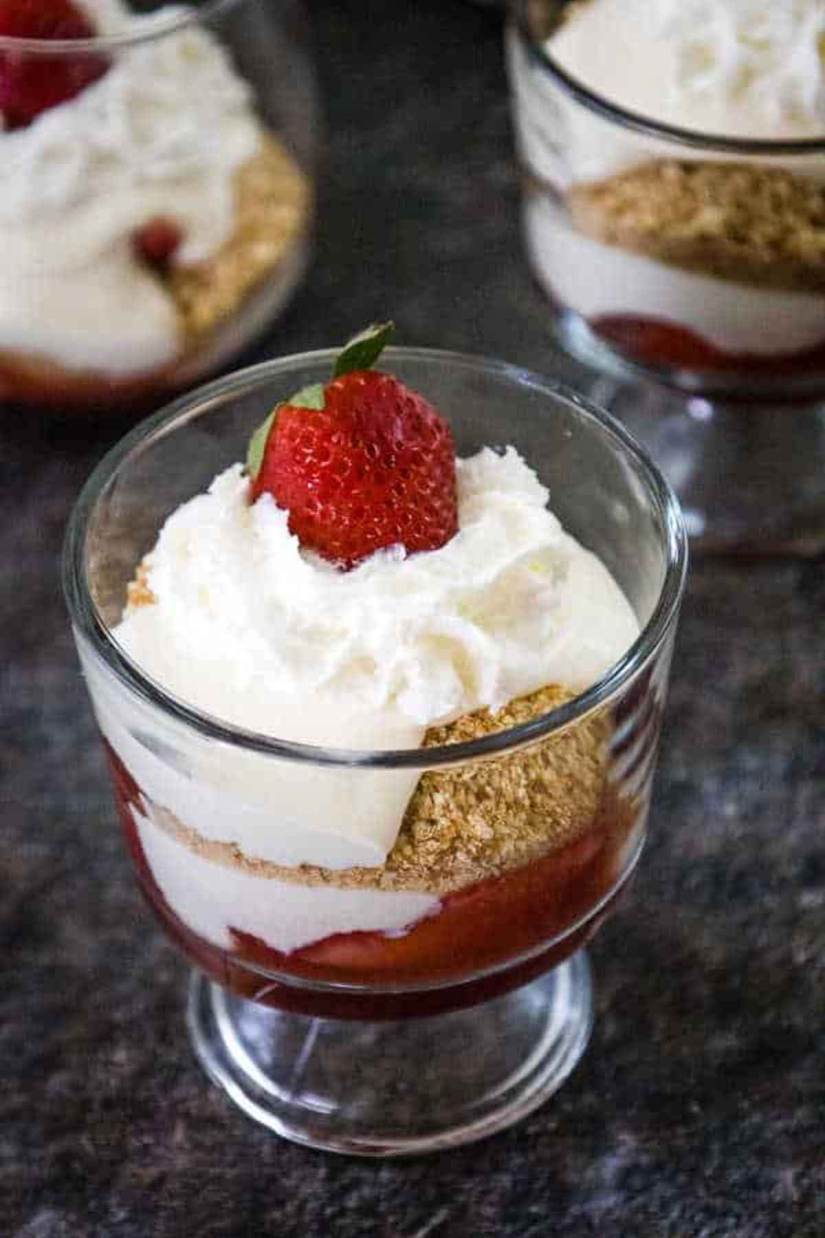 A glass dessert cup fllled with layers of strawberry sauce, creamy cheesecake filling and crushed vanilla wafer cookies topped with whipped cream and a fresh strawberry.