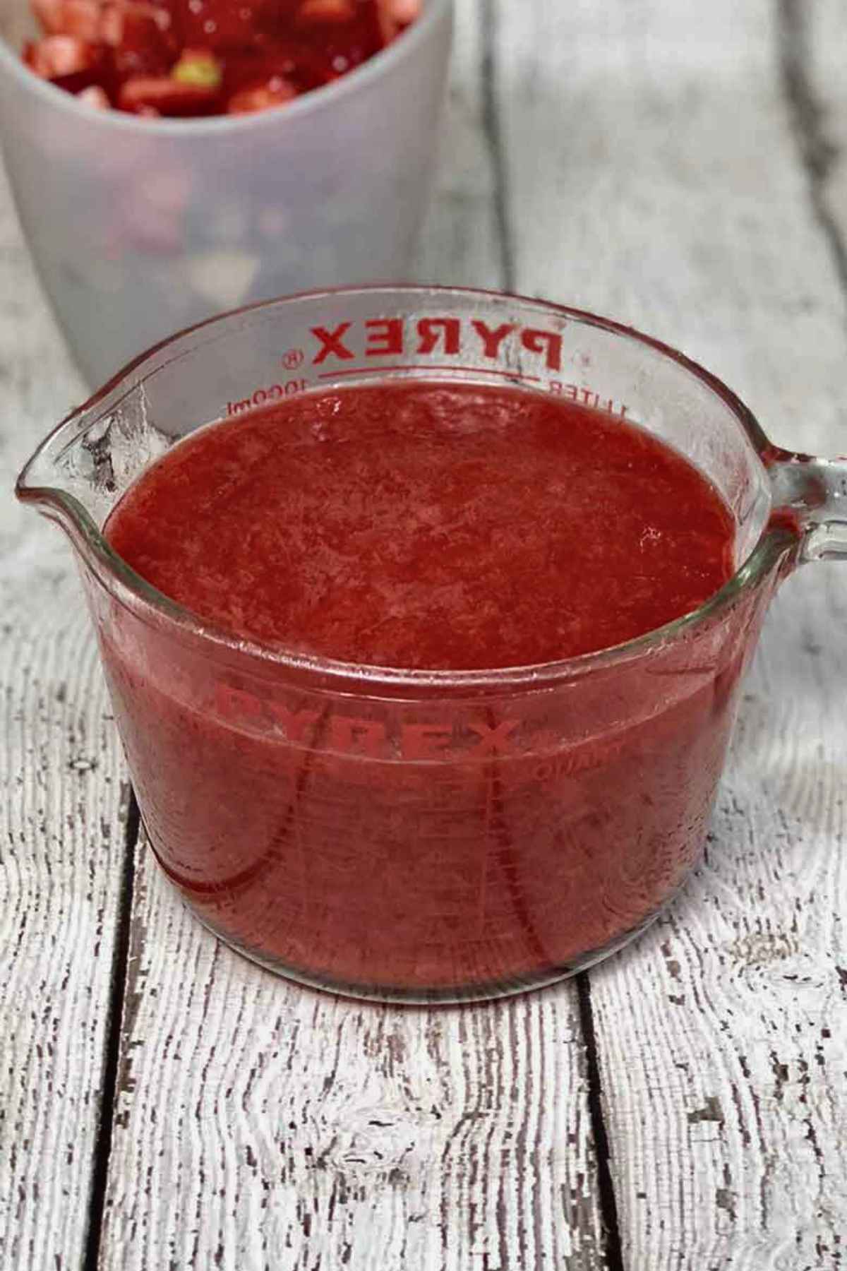 A pyrex measuring cup filled with freshly made strawberry rhubarb compote, sitting on a white wooden surface with a blurred bowl of fruit in the background.