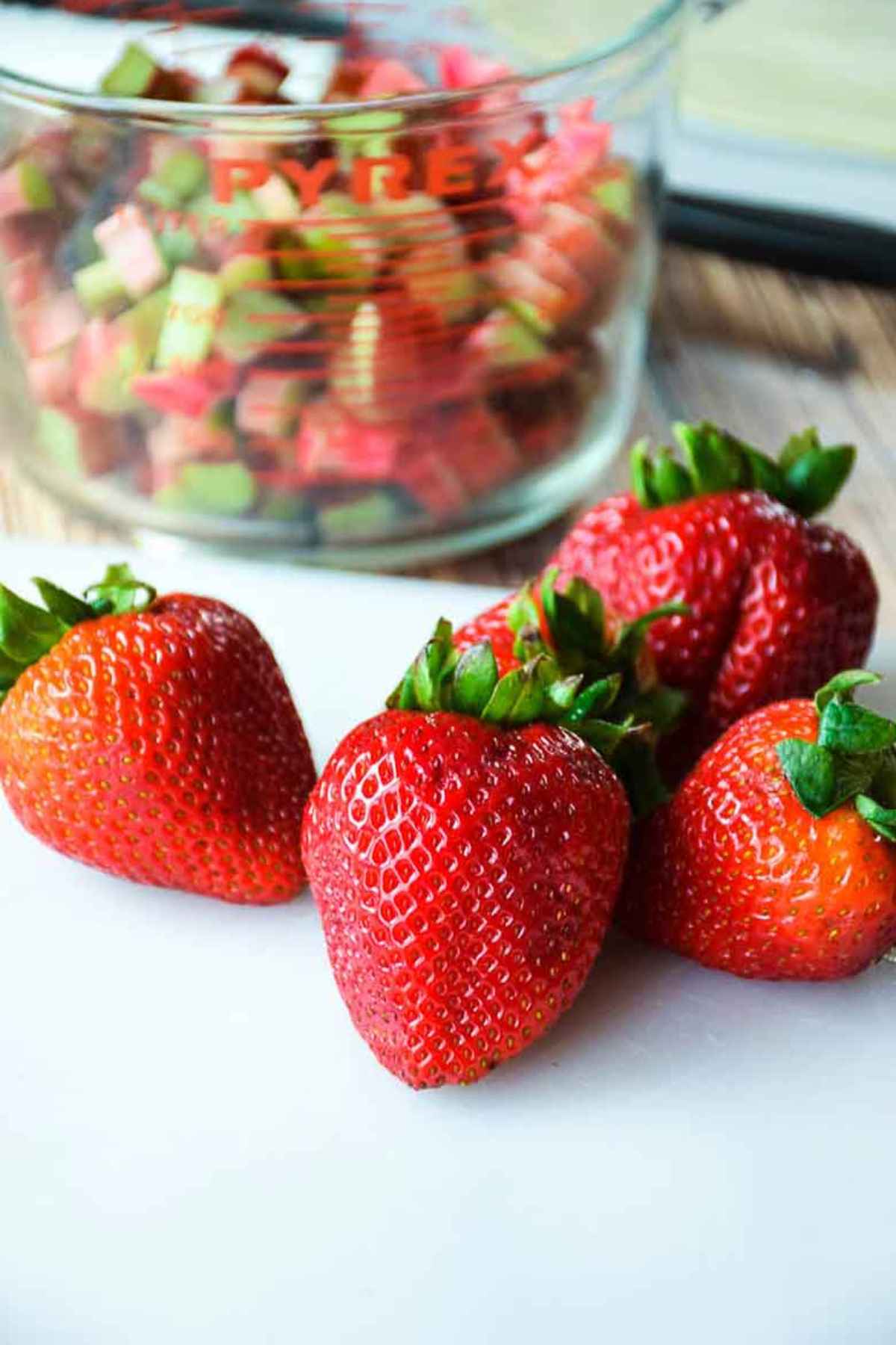 Fresh strawberries on a cutting board with a glass measuring cup of diced rhubarb in the background.
