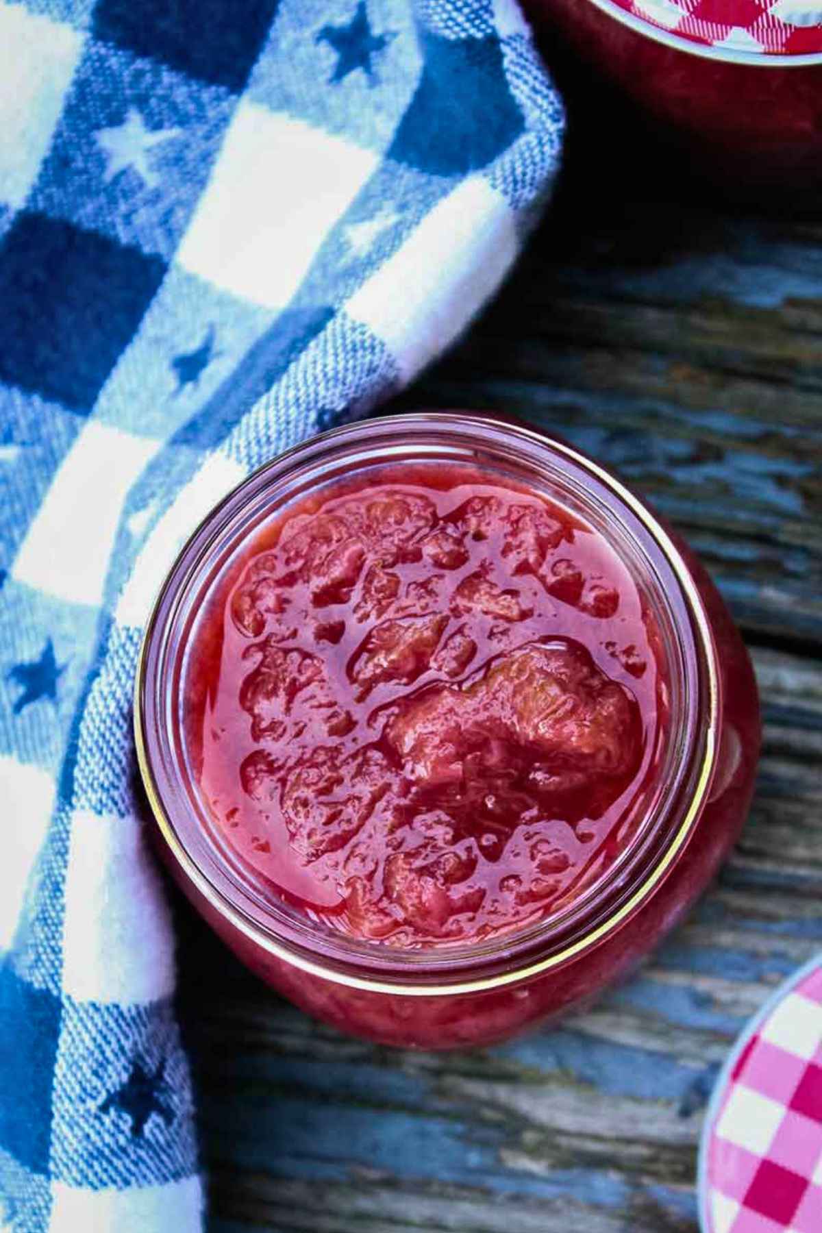 Overhead view of a jar of strawberry rhubarb compote on a rustic wooden table, next to a blue star patterned cloth and a red gingham lid.