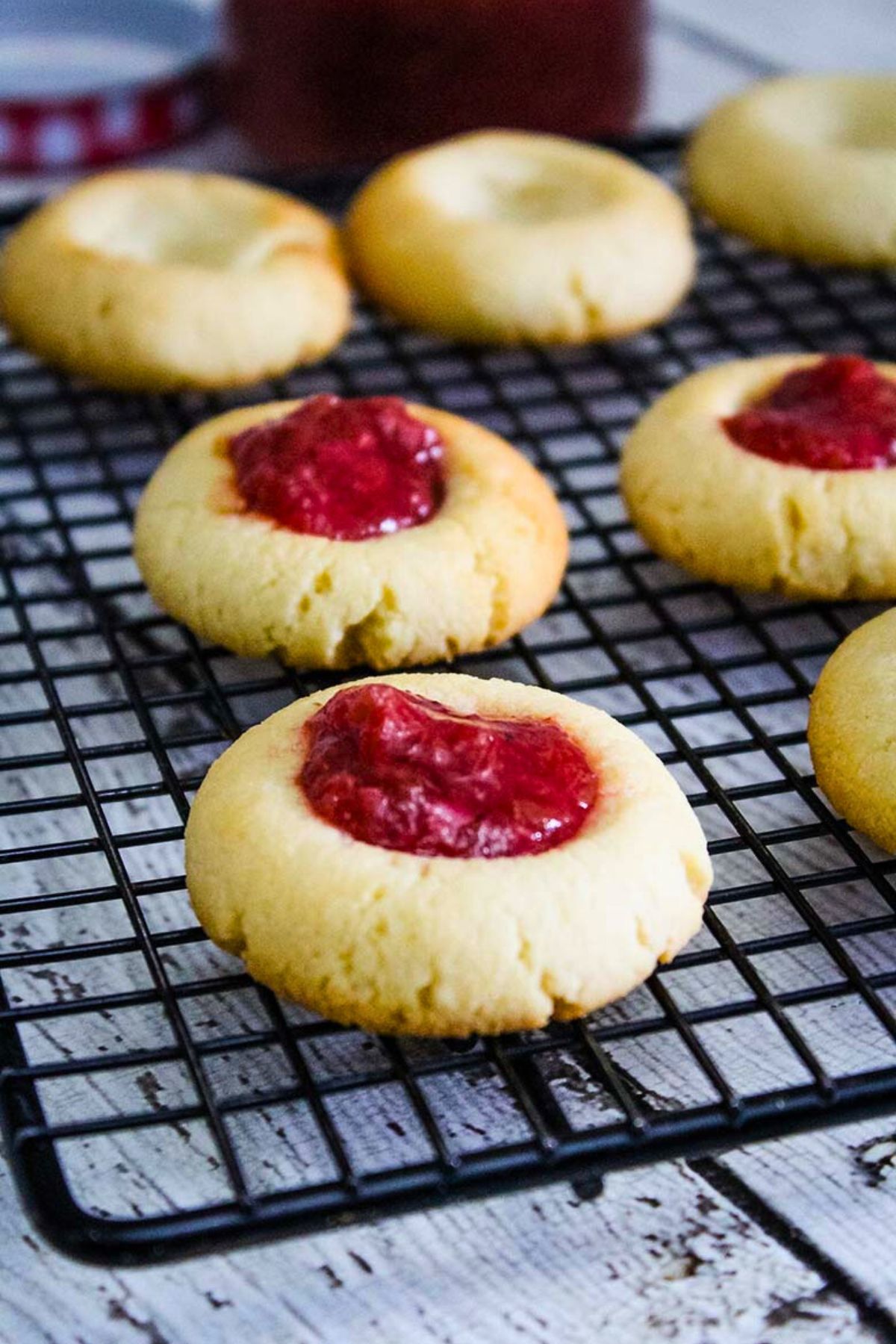 Golden thumbprint cookies on a black cooling rack, some filled with strawberry rhubarb compote and others unfilled.