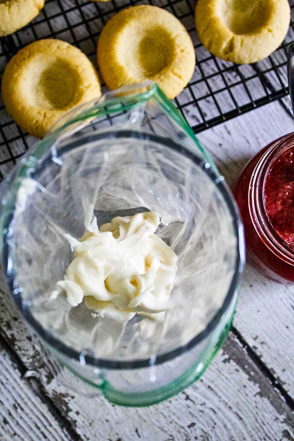 A piping bag filled with white icing next to a jar of strawberry rhubarb compote and unfilled thumbprint cookies on a black cooling rack.