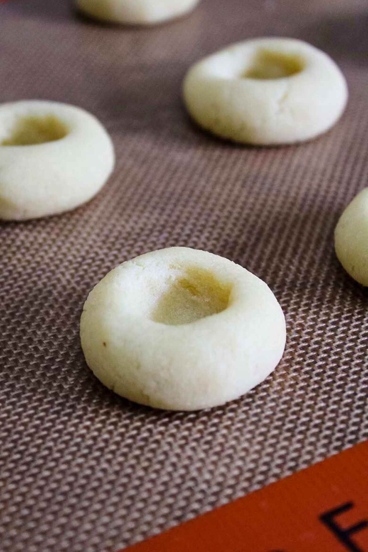 Unbaked thumbprint cookie dough rounds with indents in the center of a baking mat.
