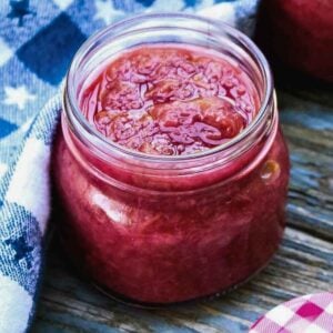 A glass jar filled with chunk strawberry rhubarb compote, placed on a rustic wooden surface next to a blue checkered cloth.
