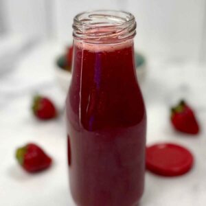 Clear glass bottle filled with homemade strawberry syrup, with fresh strawberries blurred in the background.