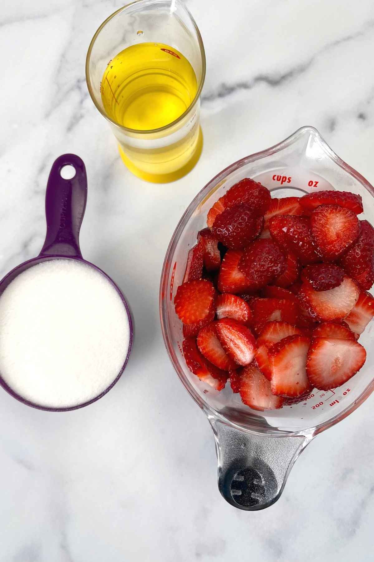Measured ingredients including sliced strawberries in a measuring cup, a cup of sugar and a cup of water on a marble surface.