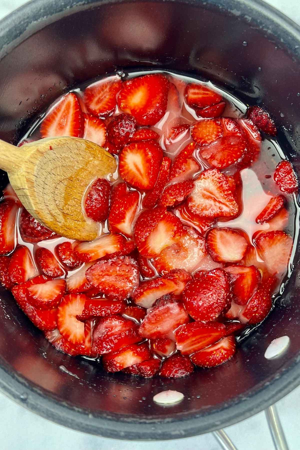 Sliced strawberries simmering in a saucepan with a wooden spoon.
