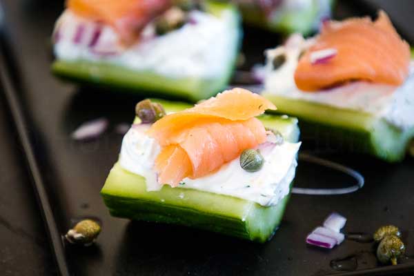 Cucumber bites topped with cream cheese, smoked salmon, capers, and red onion on a black plate.