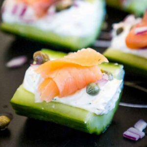 Cucumber bites topped with cream cheese, smoked salmon, capers, and red onion on a black plate.