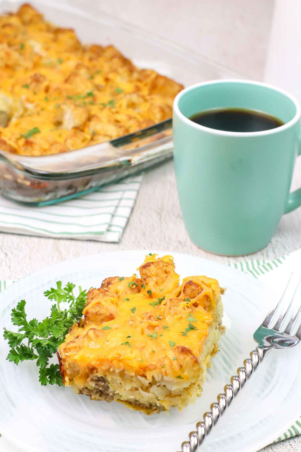 Cheesy tater tot breakfast casserole served with coffee, plated with a fork and parsley garnish.