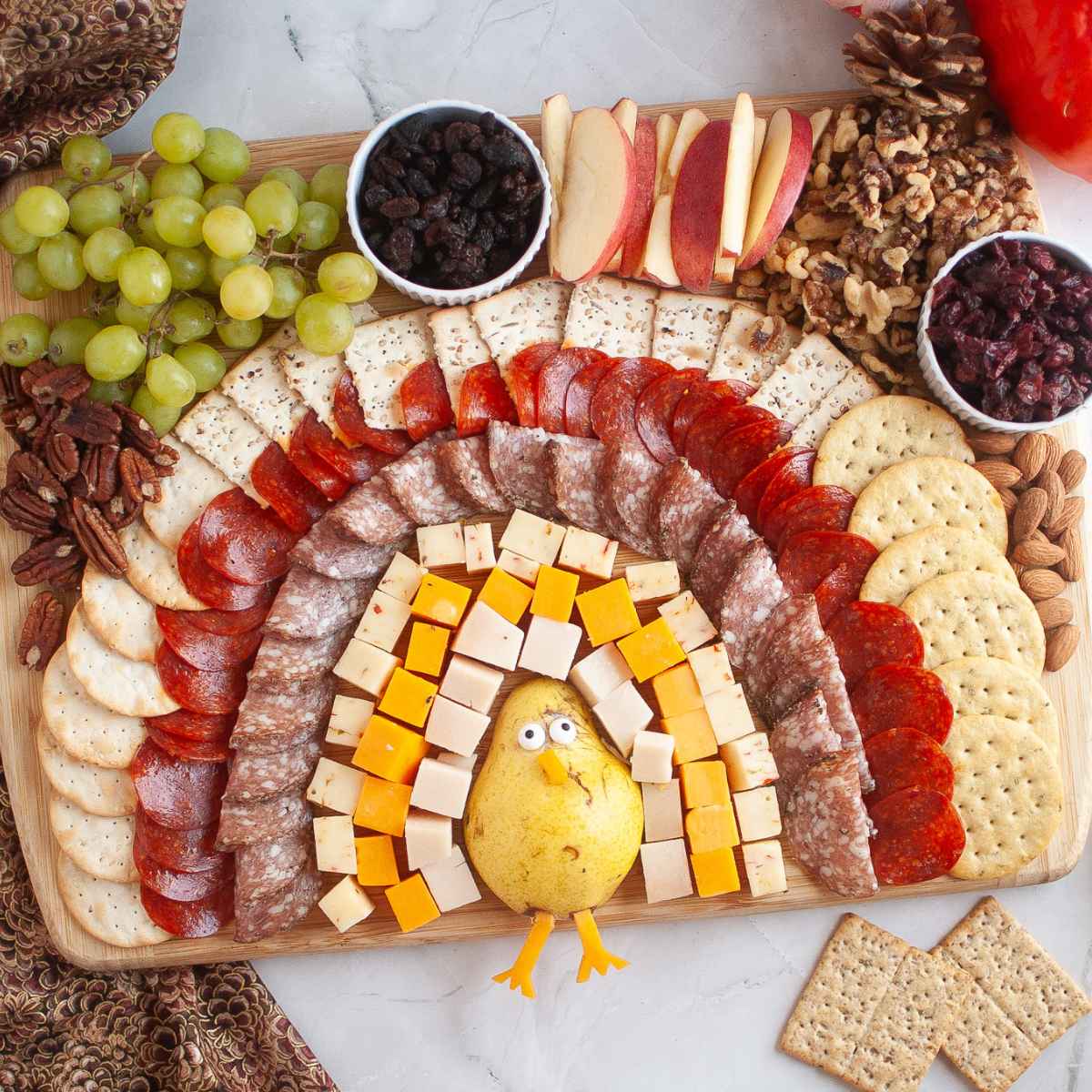 A turkey charcuterie board arranged to look like a turkey, with a pear body, cheese cubes as feathers, layers of meats, crackers, fruit, and nuts.