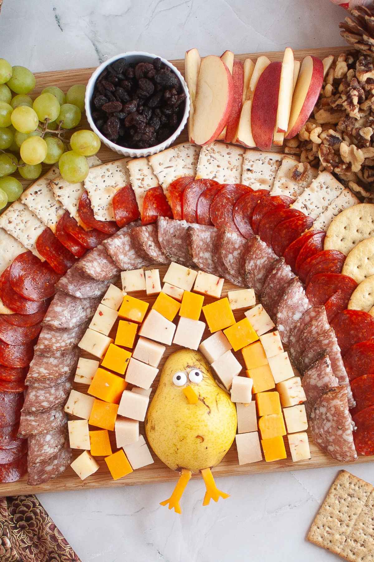 A turkey charcuterie board arranged to look like a turkey, with a pear body, cheese cubes as feathers, layers of meats, crackers, fruit, and nuts.