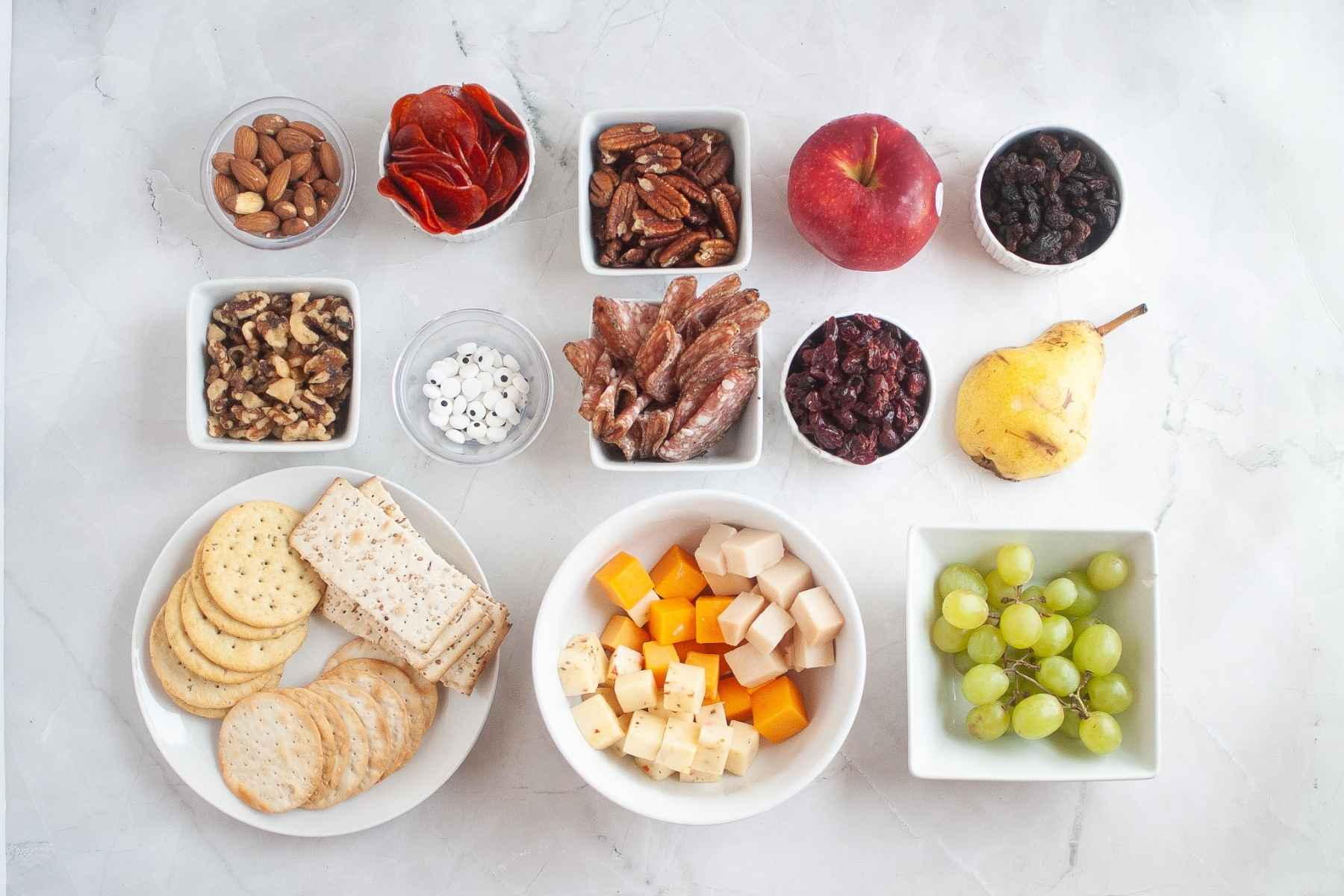 Individual ingredients for a charcuterie board laid out on a table, including cheese cubes, meats, crackers, grapes, apple, pea, nuts, and dried fruit.