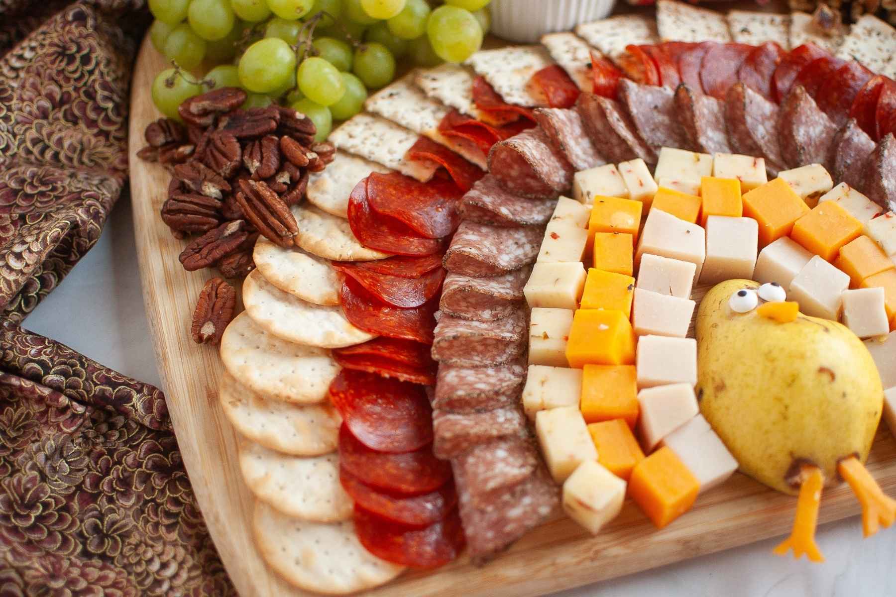 Close up horizonal view of a turkey-themed charcuterie board with colorful rows of cheese, meats, crackers, grapes, pecans, and a pear turkey centerpiece.
