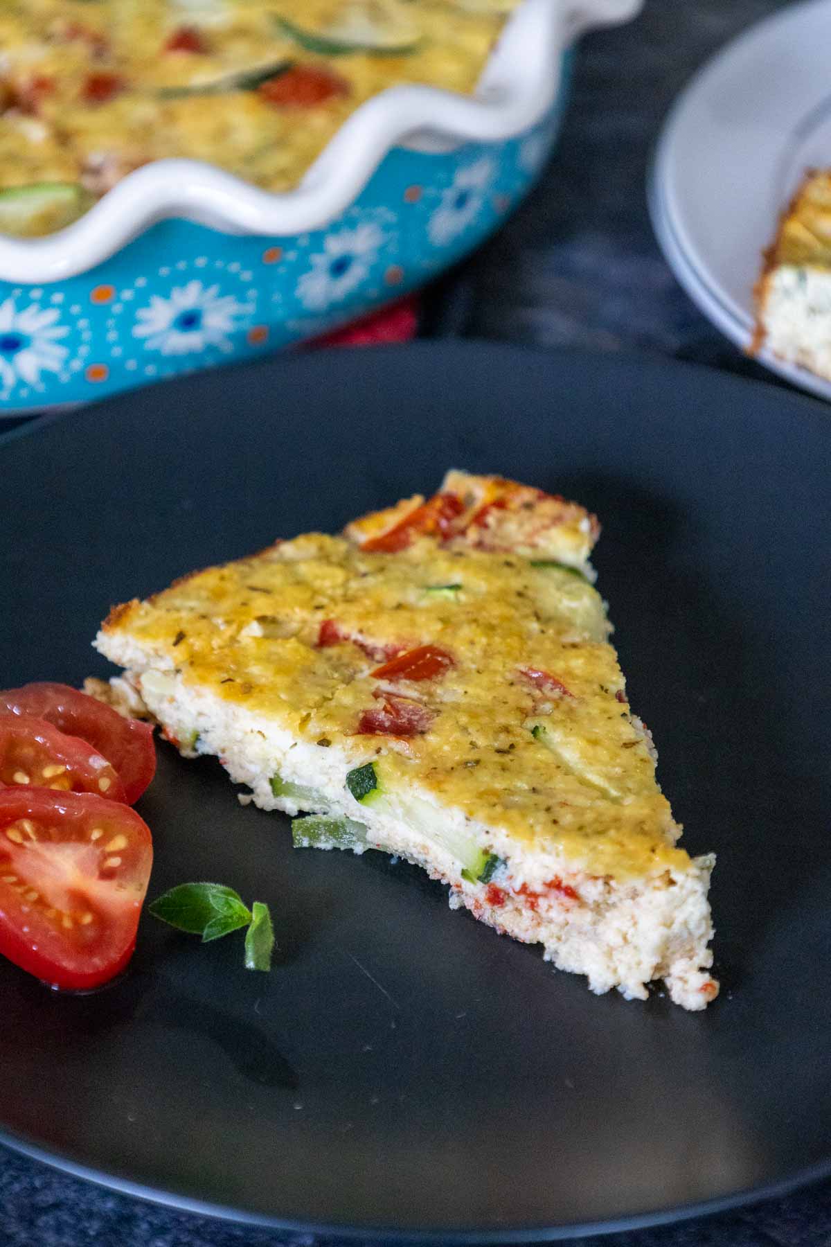 A wedge of keto zucchini pie on a black plate with a sliced cherry tomato on the side garnisheed with fresh thyme.