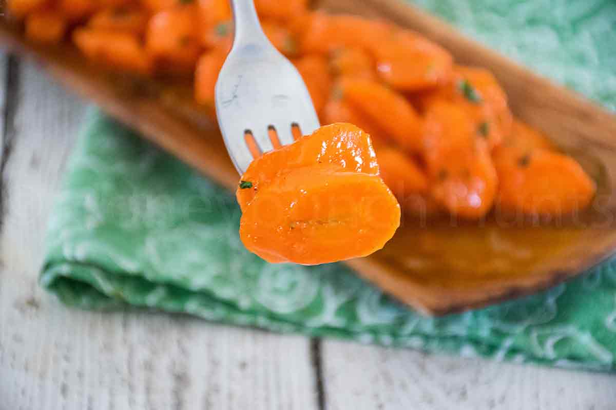Fork holding a honey glazed carrot slice with more carrots in the background.