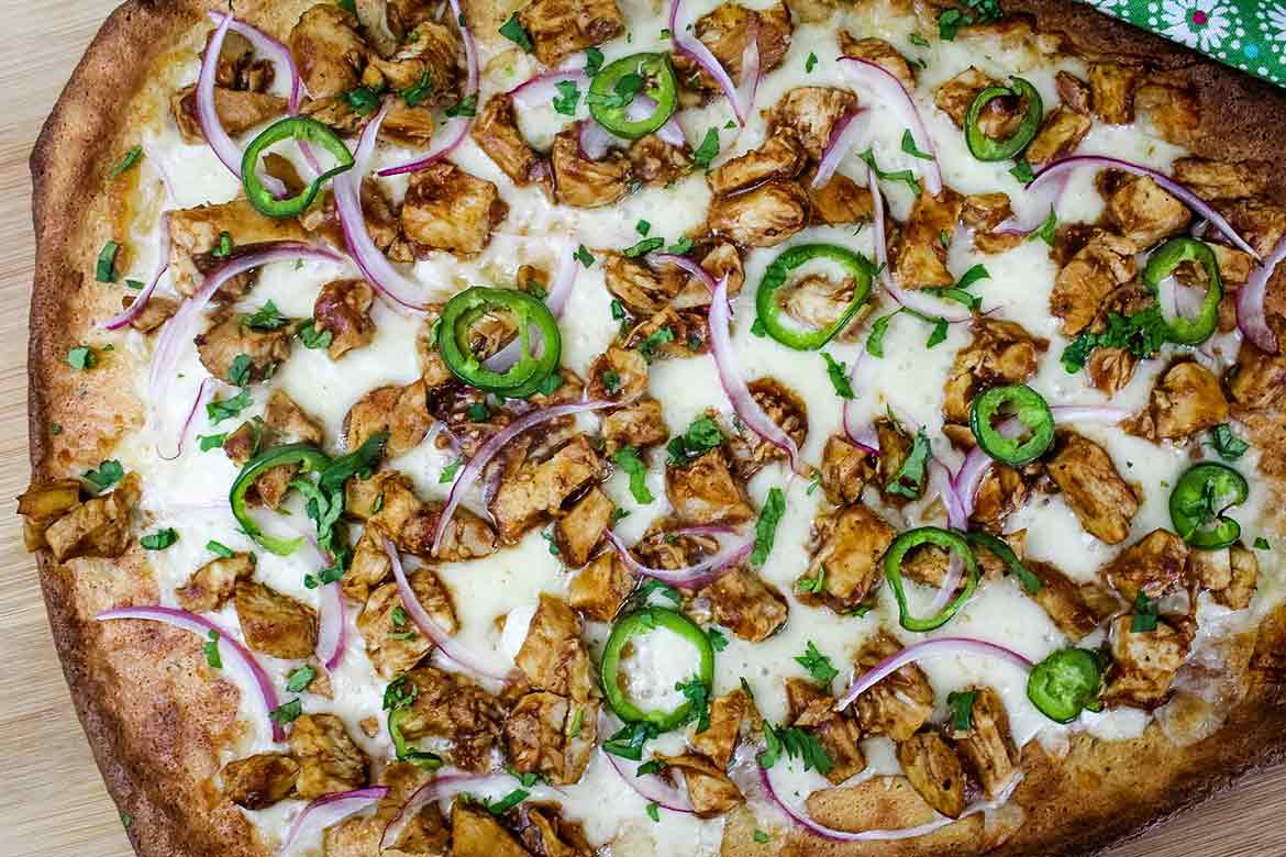 Freshly baked bbq chicken pizza topped with melted cheese, chicken, red onions, jalapenos, and cilantro, resting on a wooden board with a green patterned napkin.