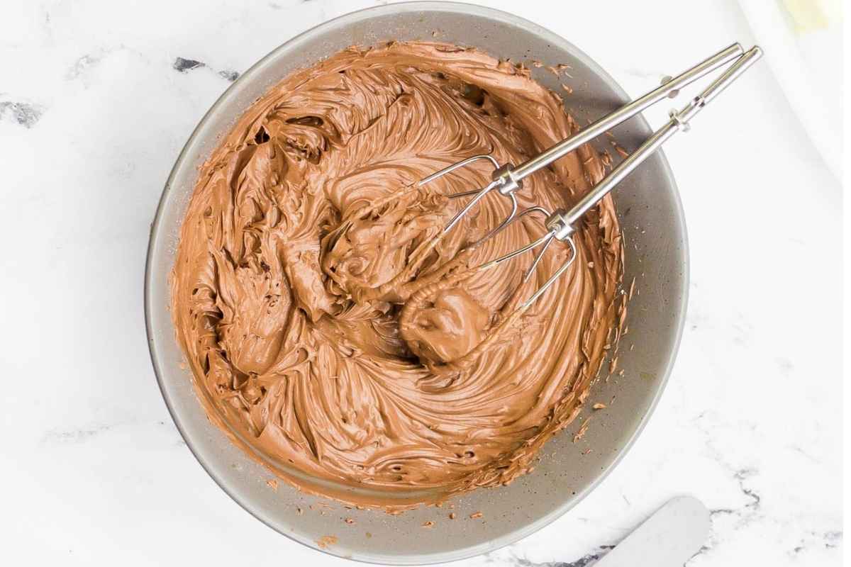 A mixing bowl filled with creamy chocolate frosting with two metal beaters resting inside.