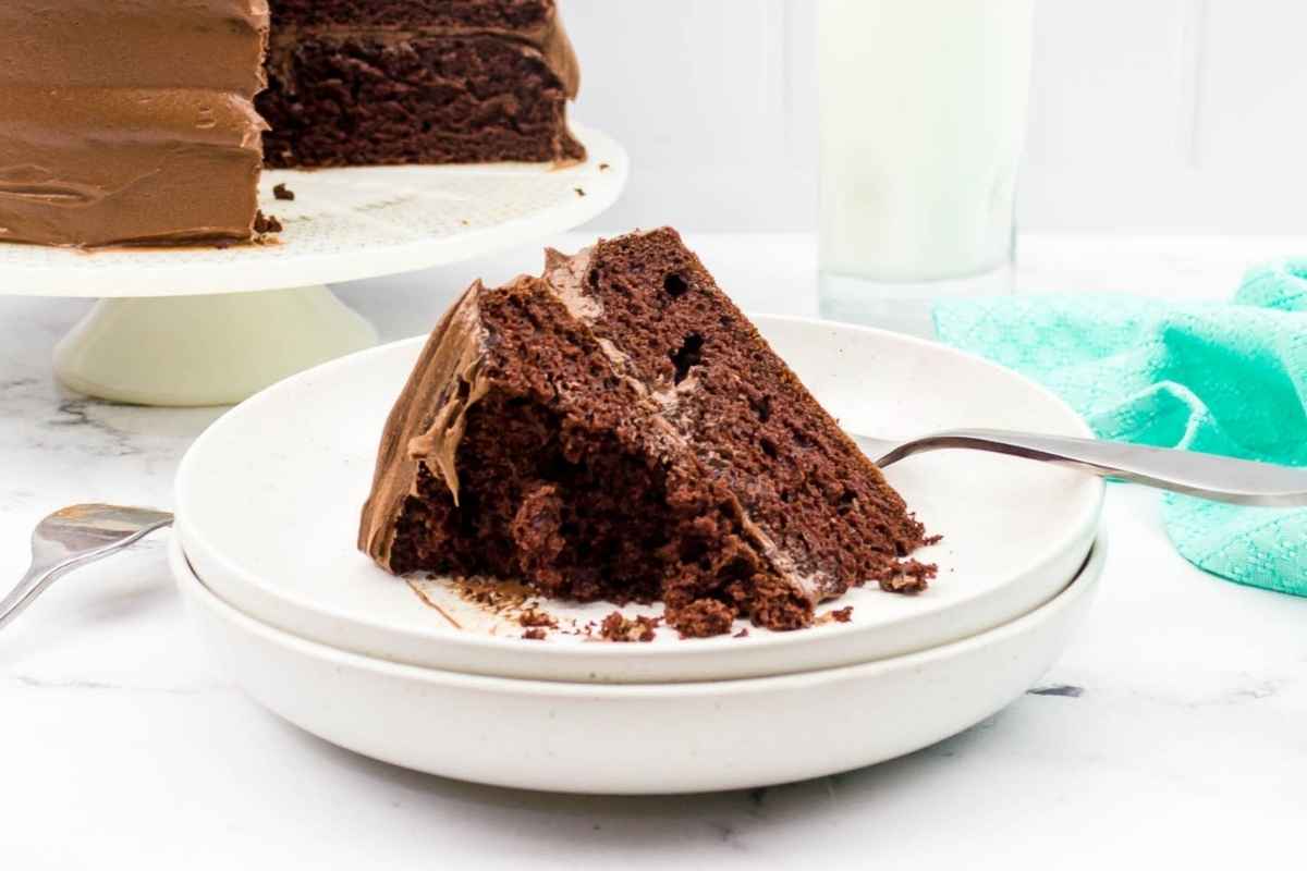 A slice of frosted chocolate layer cake on a white plate with the full cake on a stand in the background.