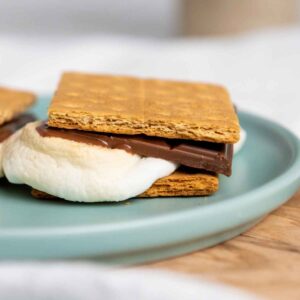 Close up side view of an Air Fryer S'more with melted chocolate and toasted marshmallow between graham crackers on a teal plate.